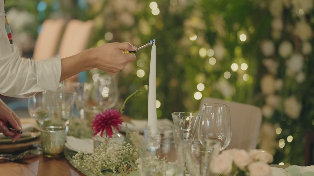 Mano che accende una candela alta su un tavolo con vetreria e fiori