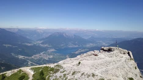 Luftaufnahme-Eines-Jungen-Mannes,-Der-Auf-Der-Spitze-Der-Cima-Vezzena,-Pizzo-Di-Levico,-Steht-Und-Eine-Drohne-Nach-Vorne-Fliegt,-Mit-Panoramablick-Auf-Trient,-Italien