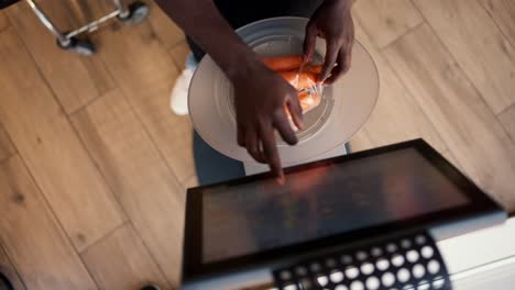 Close-up-shot-from-above:-a-Black-skinned-man-in-a-white-T-shirt-and-black-apron-selects-the-desired-product-on-the-display-of-electronic-scales-and-weighs-vegetables