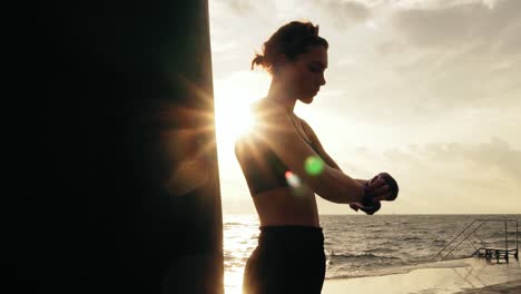 Camera-round-movement:-female-boxer-wrapping-hands-with-boxing-wraps-on-the-beach-by-the-ocean-standing-against-the-son.-Lens