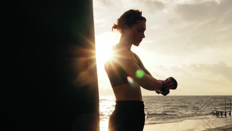 movimiento redondo de la cámara: boxeadora envolviendo las manos con envolturas de boxeo en la playa junto al océano de pie contra el hijo. lente