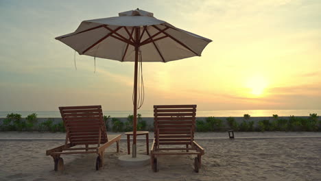 Close-up-of-two-empty-beach-chairs-and-a-shade-umbrella-on-a-sandy-beach-facing-a-colorful-ocean-sunset