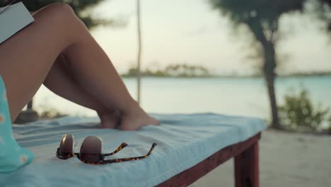 Close-up-view-of-woman-laying-on-a-sunbed,-reading-a-book-and-drinking-from-a-coconut