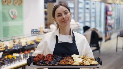 A-happy-worker-woman-in-a-black-apron-is-holding-a-crate-of-cakes.-Work-in-the-store.-Weekdays