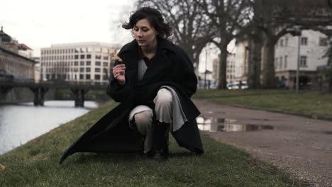 Young-woman-in-a-black-coat-standing-in-a-park-in-autumn-in-afternoon-and-pick-up-a-leaf