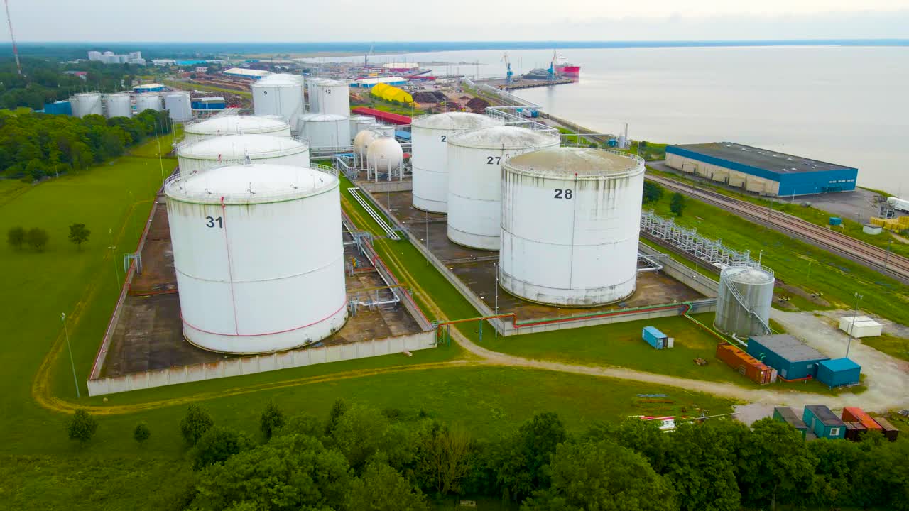 Aerial drone footage orbiting around large white colored bulk liquid storage vessles or containers that are by Paldiski seaside industrial shoreline with a container trading port or harbor visible