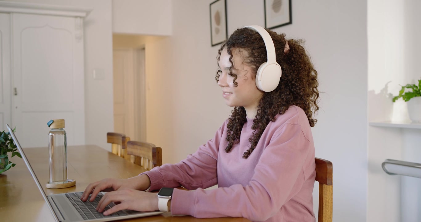 Teenage woman of African descent studying, typing on laptop at dining table wearing headphones