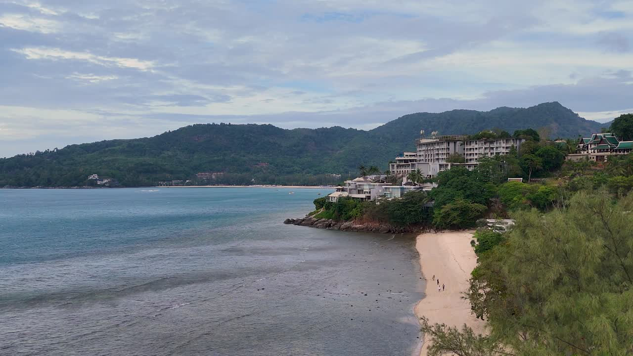 las imágenes de drones capturan una serena costa de phuket con exuberante vegetación y edificios turísticos bajo un cielo nublado