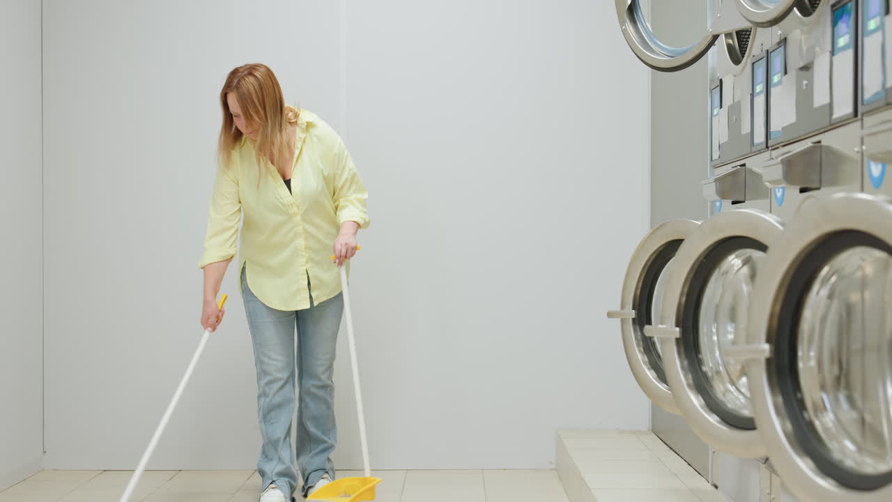 Zoom out shows elegant woman near industrial washing machine in laundromat, yellow shirt and jeans with white sneakers sweeping tiled floor using broom and dustpan, cleaning scene
