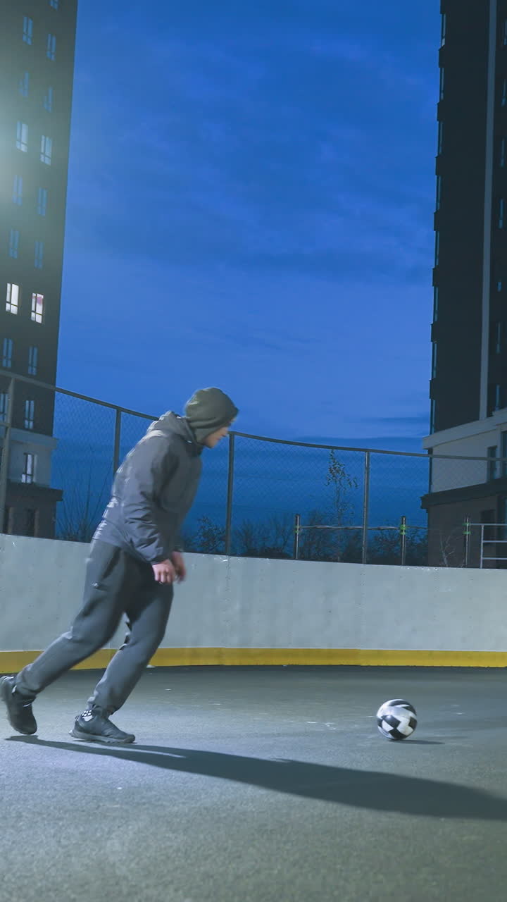 jugador de fútbol patea la pelota de fútbol con precisión hacia la meta durante la sesión de entrenamiento nocturna en un campo urbano iluminado, rodeado de altos edificios residenciales y un vibrante cielo azul de la tarde