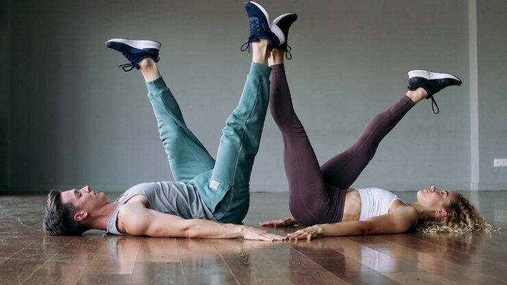 Starting bent knees, pair doing leg lifts on hardwood floor for core, palms touching, in sportswear