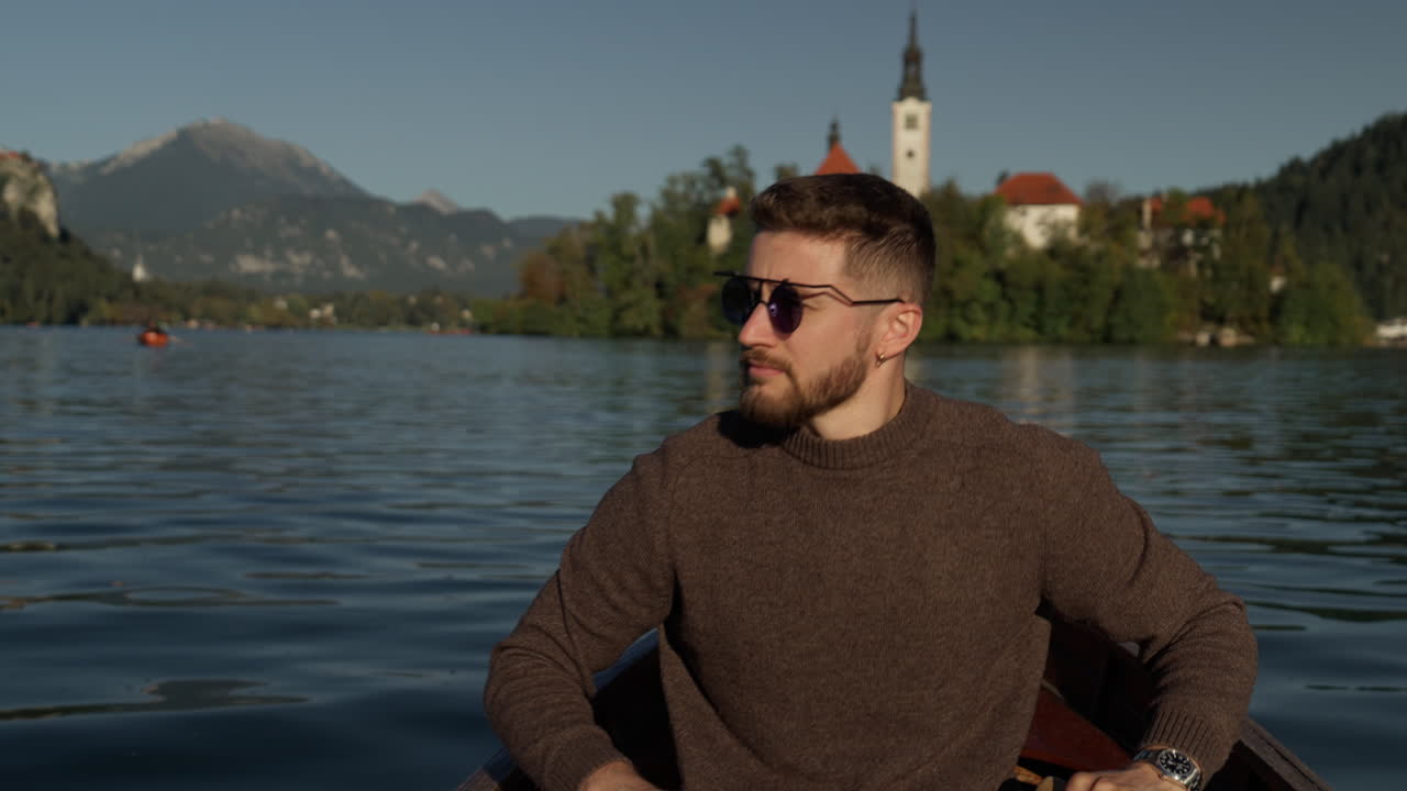 Autumn cinematic footage of a man rowing a wooden boat on Lake Bled, Slovenia. The church on the island is visible in the background under sunny skies