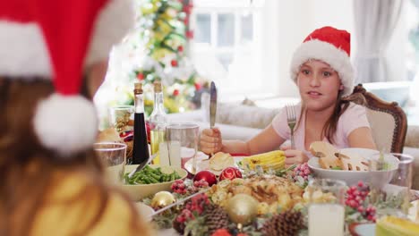 chica caucásica en sombrero de santa con tenedor y cuchillo mientras está sentada en la mesa de comedor y lista para disfrutar