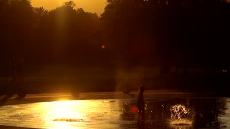 Niño-Pequeño-Jugando-Con-Fuente-De-Agua-Contra-Un-Fondo-De-Puesta-De-Sol-En-Cámara-Lenta