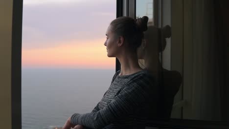 close up view of beautiful young woman standing by the open window during the sunset by the sea and enjoying the view. beautiful