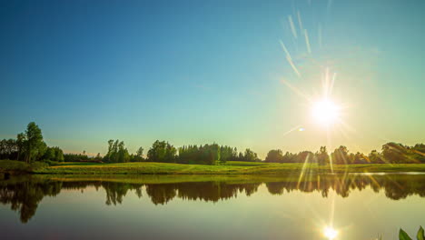 Timelapse-shot-of-a-reflective-lake-along-green-grasslands-and-trees-with-sun-setting-in-the-background