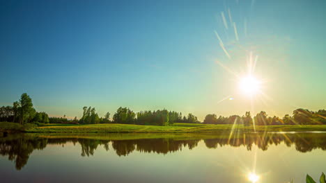 timelapse shot of a reflective lake along green grasslands and trees with sun setting in the background