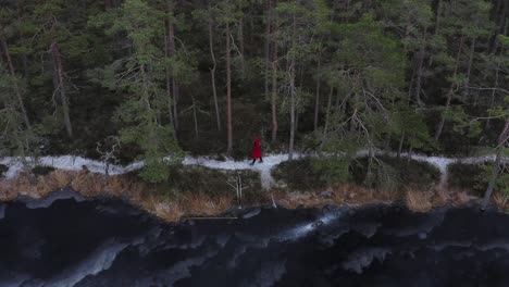 Woman-Wearing-Red-Hoodie-Coat-In-Snowy-Lane-By-The-Freezing-Lake-During-Winter