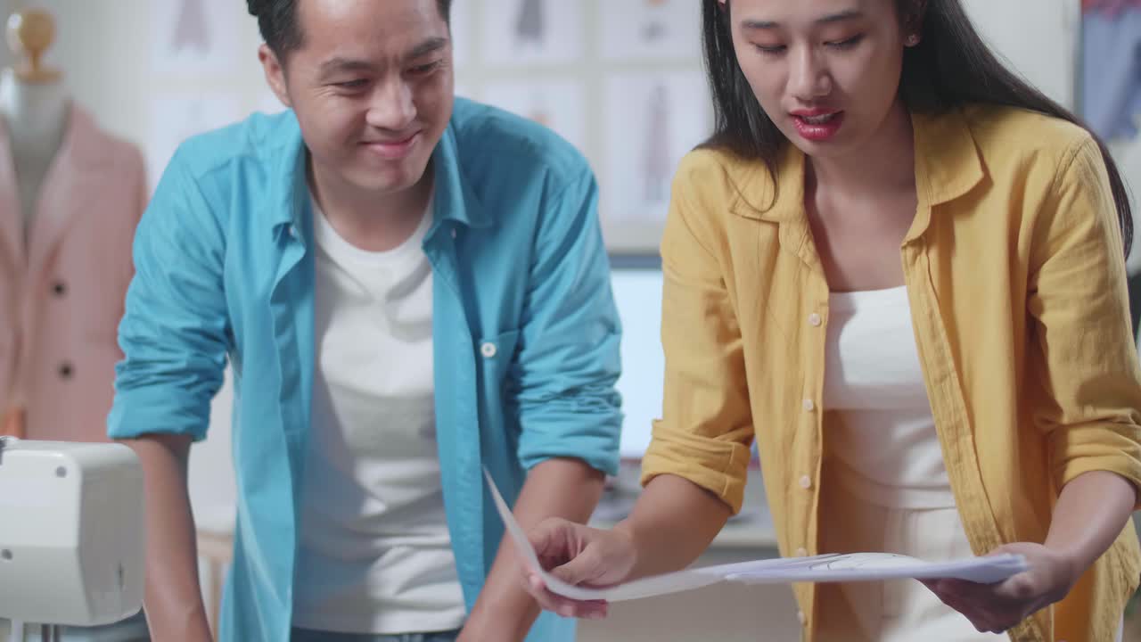 아시아의 남녀 디자이너들이 바느질기를 들고 스튜디오에서 옷을 디자인하는 동안 서로 서류를 재배열하는 데 도움을 주고 있습니다.