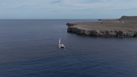 single catamaran boat sails off coast of pont den gil, minorca spain at midday with sail up