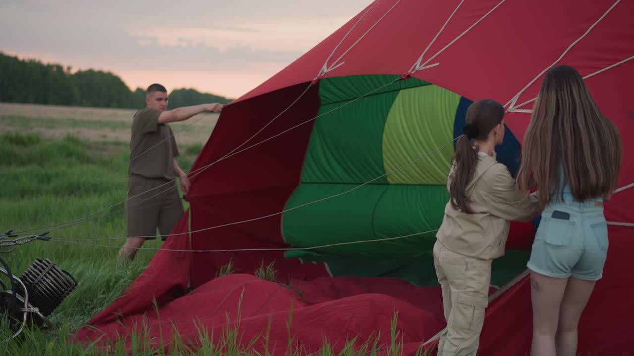 family prepping hot air balloon in open field at sunset with teamwork as woman in brown outfit assists young lady holding vibrant envelope while man in olive attire steadies fabric inflating envelope