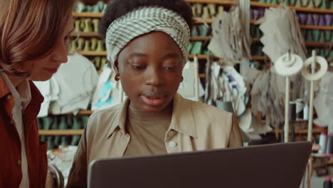 mujeres alegres usando computadora portátil y charlando en un taller de fabricación de zapatos