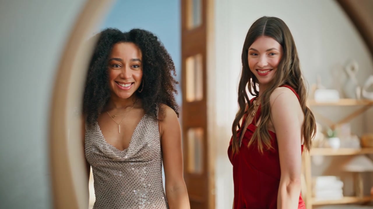 Duas mulheres deslumbrantes usando vestidos elegantes verificando o visual no espelho em close-up.