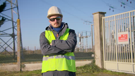 ingeniero masculino con chaleco reflectante de seguridad y gafas de sol mientras cruza los brazos en la subestación eléctrica, dinámica de mano