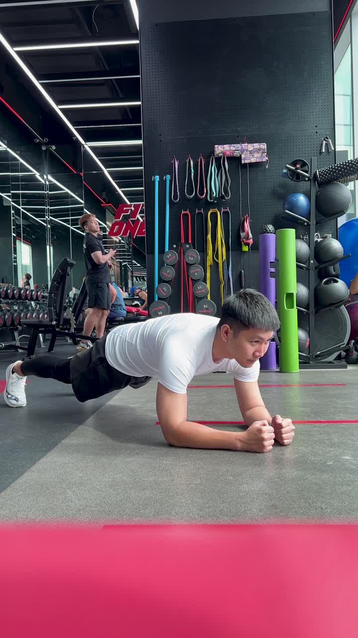 Una persona realiza un ejercicio de plancha sobre una colchoneta de gimnasio, rodeada de equipo de fitness en un entorno de gimnasio bien iluminado.