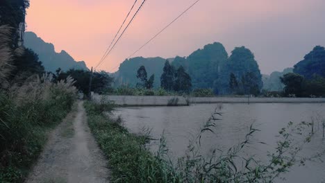 Cielos-Anaranjados-Al-Atardecer-Con-Camino-Vacío-Al-Lado-De-Aguas-De-Humedales-Con-Silueta-De-Imponentes-Acantilados-En-El-Fondo-En-Ninh-Binh,-Vietnam