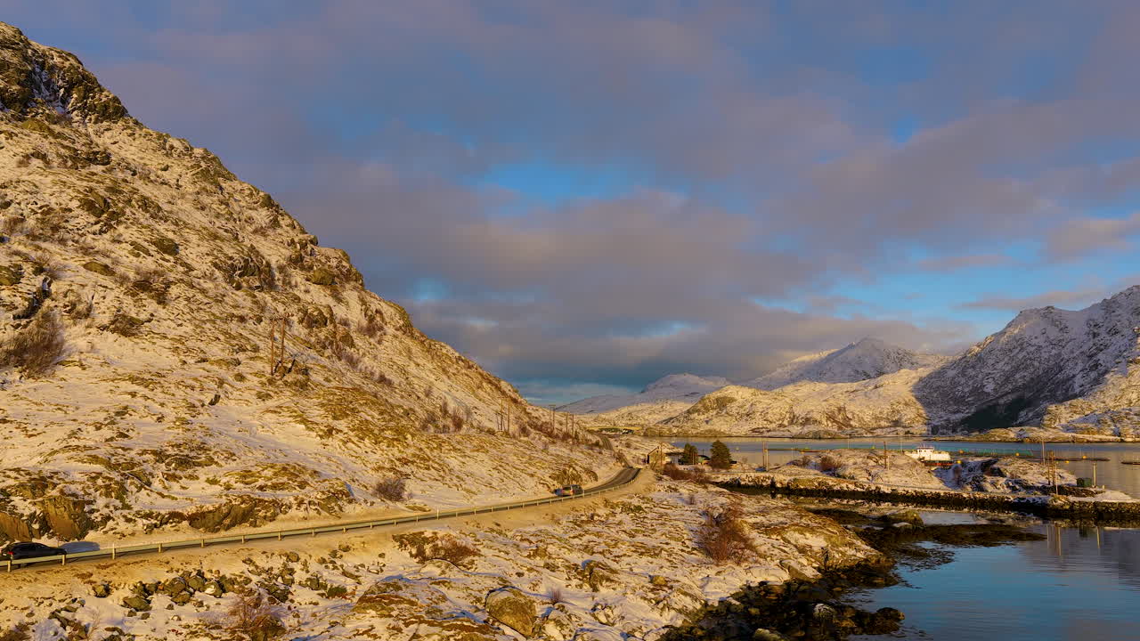 Un'inquadratura dall'alto che cattura un veicolo in viaggio lungo una strada tortuosa attraverso montagne innevate e fiordi a Lofoten, Norvegia, durante l'ora d'oro