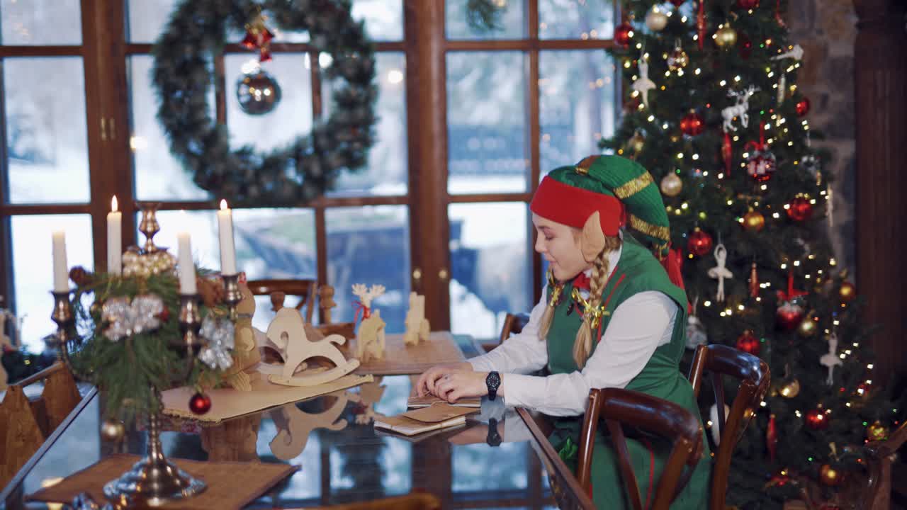 Elf with envelopes sitting at the festive table. Side view of a joyful elf opening envelope on the Christmas tree background.