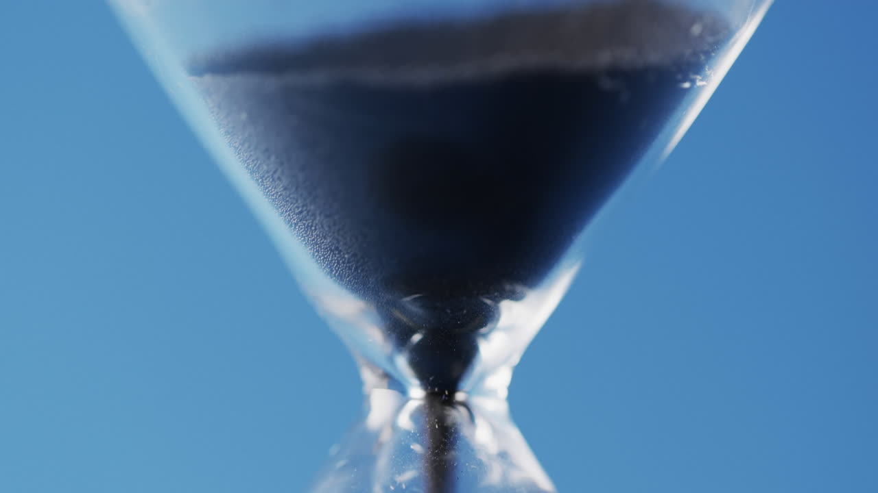 Video of close up of hourglass with black sand pouring with copy space on blue background