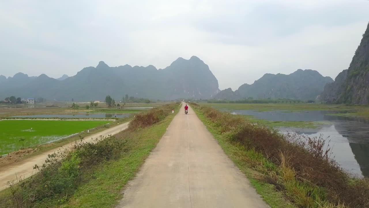 Low drone follow cam of motor biker in red driving on narrow road surrounded by grass, rice fields and lake with tall mountains in background