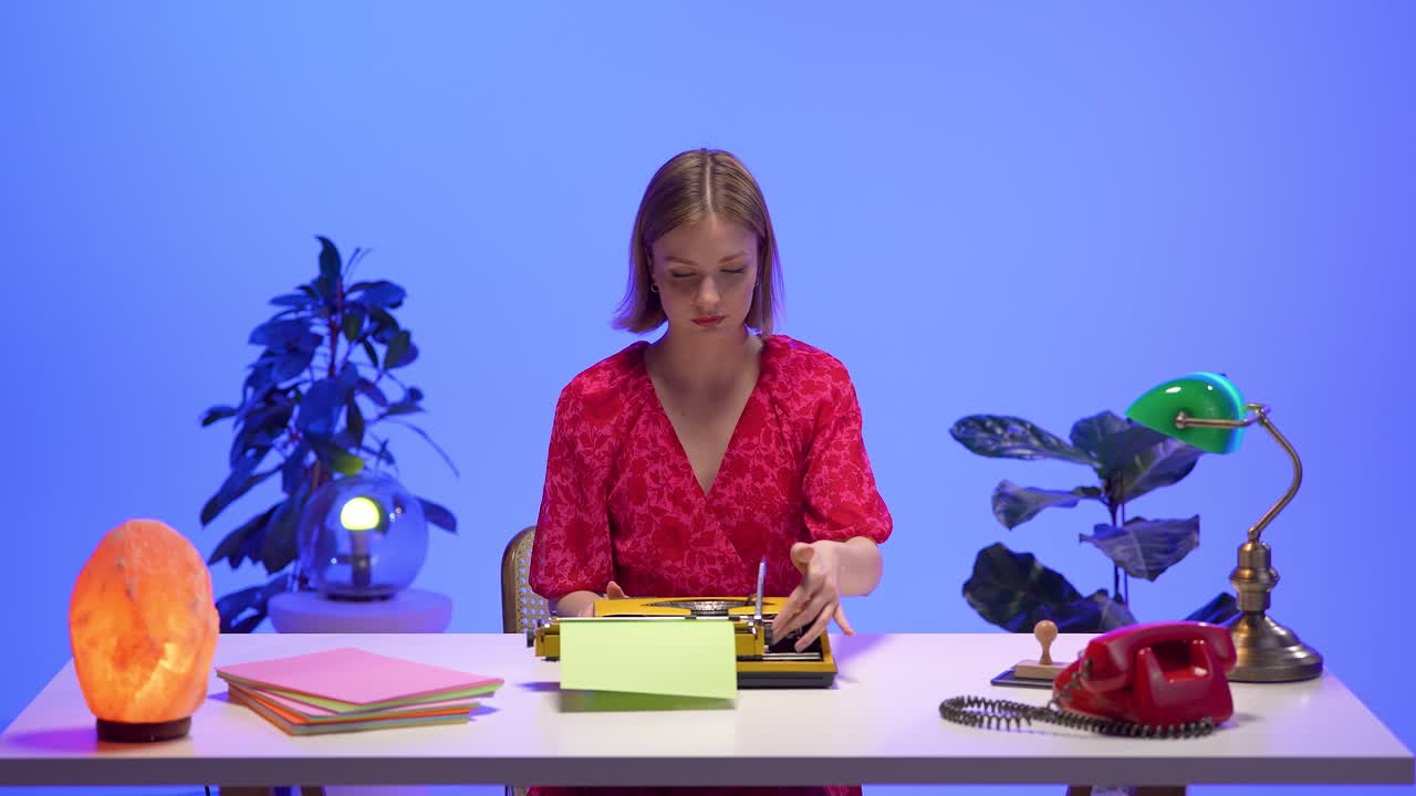 A smiling woman in a red floral dress sits behind a yellow typewriter at a retro desk. Surrounded by nostalgic props and warm lighting, she radiates calm and creativity in a vintage workspace