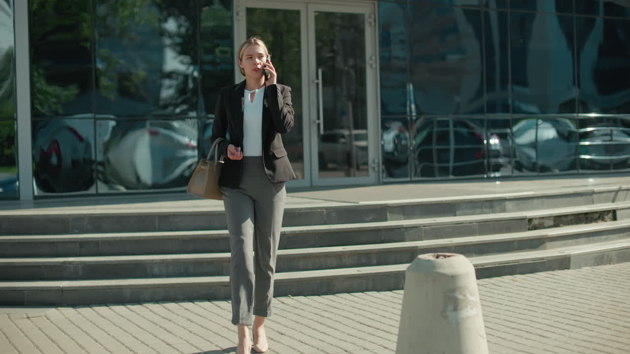 Businesswoman walking out of modern office building while talking on phone holding handbag, glass facade reflecting parked cars, metal railing along steps, sunny day, concrete bollards in foreground