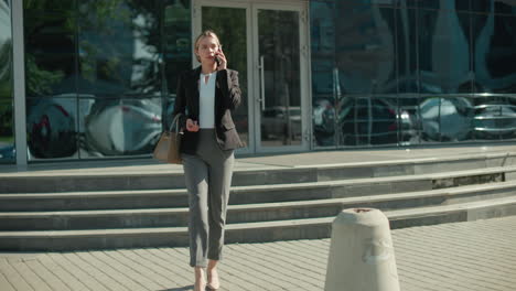 businesswoman walking out of modern office building while talking on phone holding handbag, glass facade reflecting parked cars, metal railing along steps, sunny day, concrete bollards in foreground