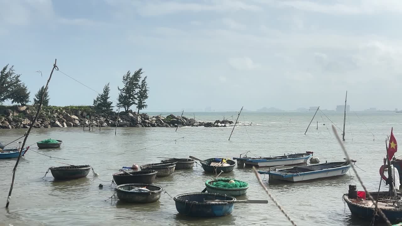 Traditionelle runde Korbboote treiben in ruhigem Wasser nahe einer felsigen Mole an der Son Tra Marina in Da Nang, während sanfte Wellen unter klarem Tageshimmel auf die Küste zurollen.