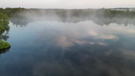 morning fog rises off reflective surface of northern lake, flyover