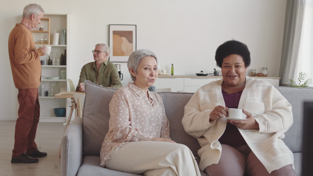 Medium long of Asian and African senior women talking, sitting on couch, watching TV, smiling, two older men communicating on background