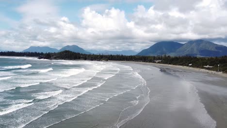 a drone video of the beach during a sunny day with large waves in the water and a forest and mountains in the backrgound