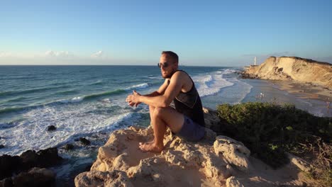 a man in sports wear and black sunglass sitting in rocky beach and looking at the blue ocean in haderah, israel