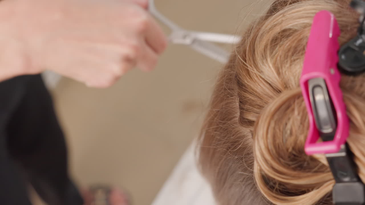 primer plano de un moño con un clip que muestra el corte preciso por parte del ayudante del estilista; el cortador sénior recortando los cabellos sueltos y rematando los bordes; suave separación con la yema de los dedos; iluminación tenue del salón; espacio de trabajo ordenado; silencio