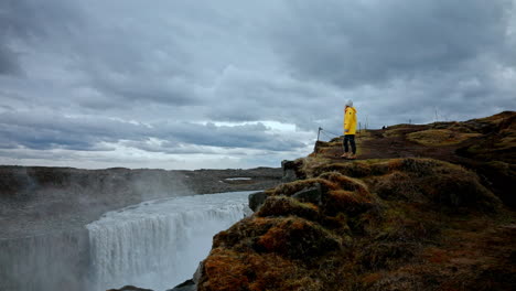 Wide-panoramic-shot-of-female-traveler-wearing-a-yellow-standing-on-the-edge-of-the-cliff-enjoying-the-view-of-the-iconic-Dettifoss-waterfall-in-Iceland