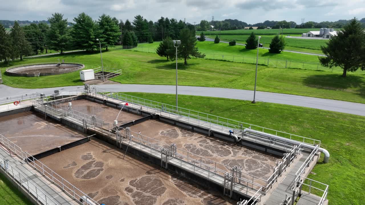 Toma aérea con inclinación ascendente de una planta industrial estadounidense de tratamiento de aguas residuales en Lititz, Pensilvania. Día soleado con césped verde en un distrito suburbano. Agua marrón en el estanque.
