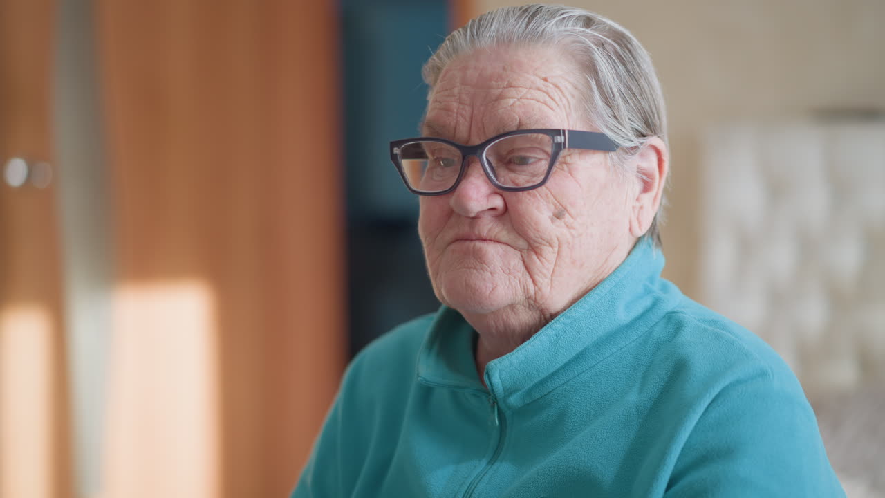 Elderly woman in a teal green sweater carefully adjusting her glasses while sitting on a bed in a bright room, focused on reading or preparing to wear them, in a serene atmosphere