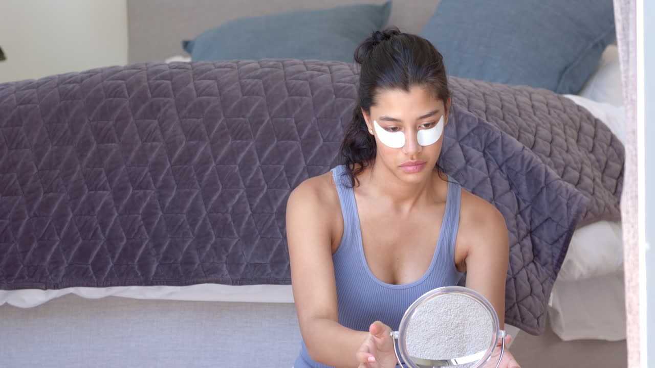 Biracial teenage girl applying under eye masks in bedroom looking in mirror, copy space, slow motion
