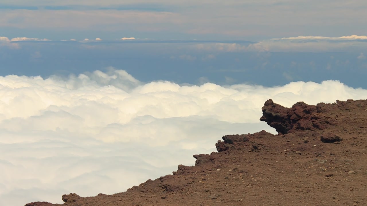 Sweeping static footage over the volcanic summit in Haleakala National Park, Maui, Hawaii, reveals a rugged rocky ridge above a thick blanket of rolling clouds beneath a tranquil blue sky.