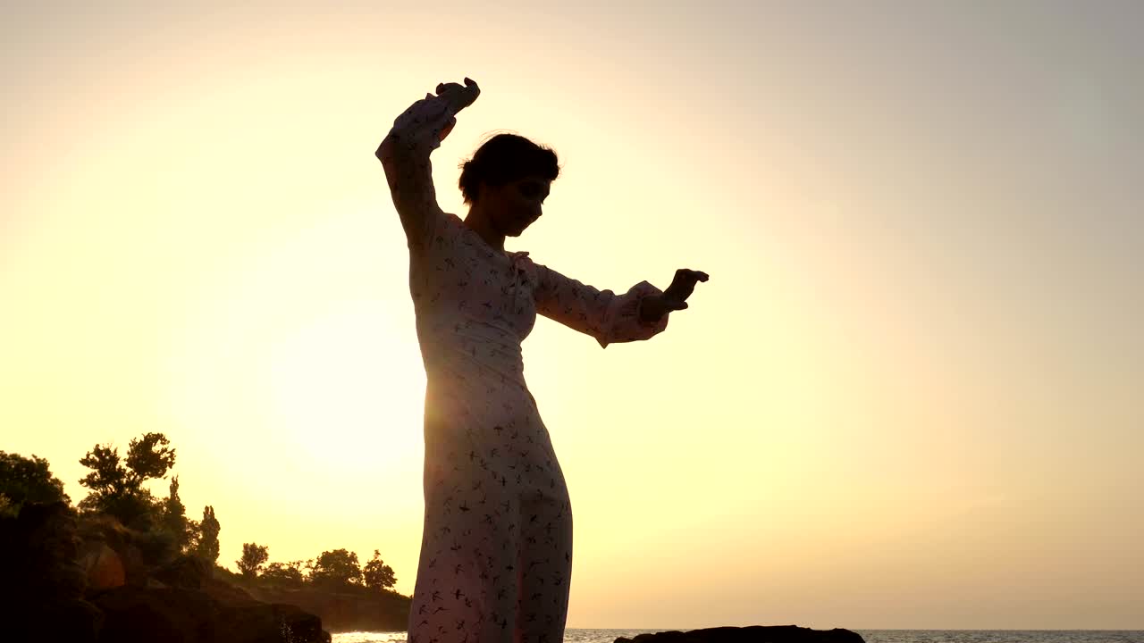 adorable chica caucásica morena en vestido claro bailando en el acantilado sobre el mar a la hora del amanecer