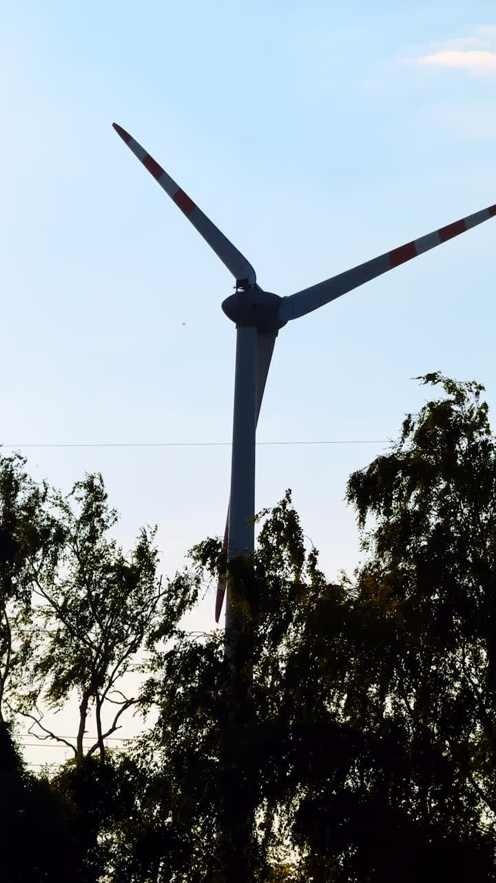 Turbine spins at sunset by trees. A wind turbine rotates against the backdrop of a colorful sunset, framed by lush trees in a tranquil setting